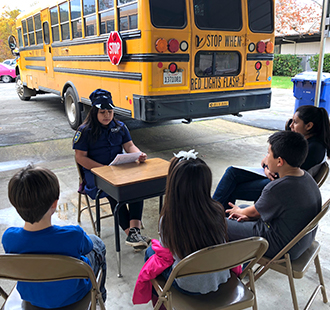Student sits at a desk outside with other students sitting in chairs in front of a school bus